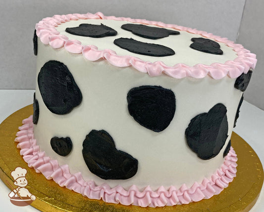 Round cake with smooth white icing and black buttercream cow print decoration with light pink ruffle trims on both the bottom and top of cake.