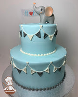2-tier round cake with plastic elephant toy sitting on top of the cake. Light blue icing and buttercream flags banner on cake walls.