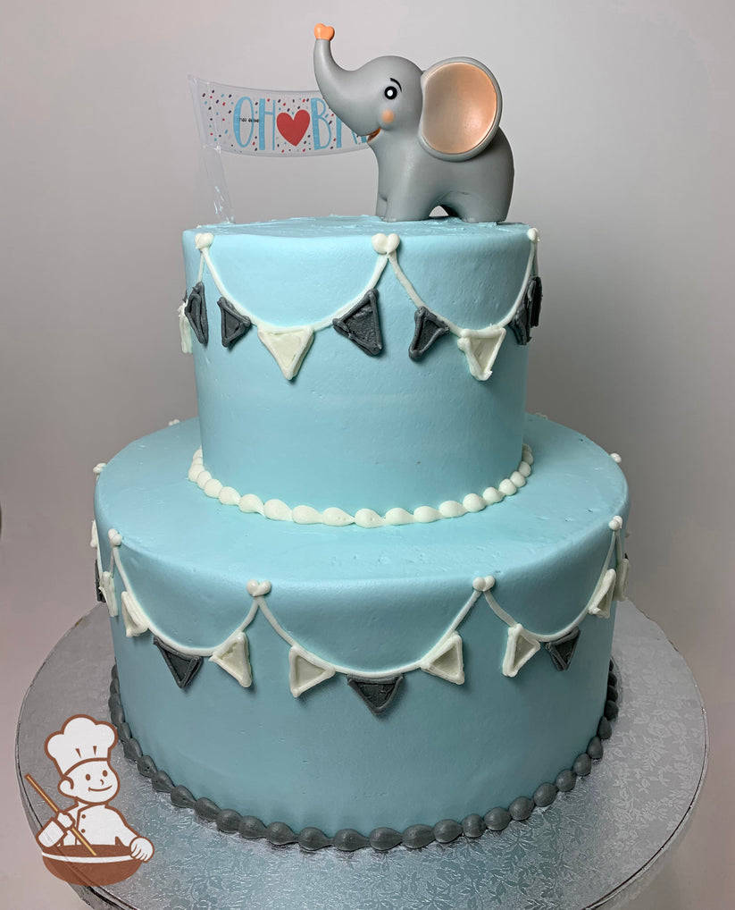 2-tier round cake with plastic elephant toy sitting on top of the cake. Light blue icing and buttercream flags banner on cake walls.