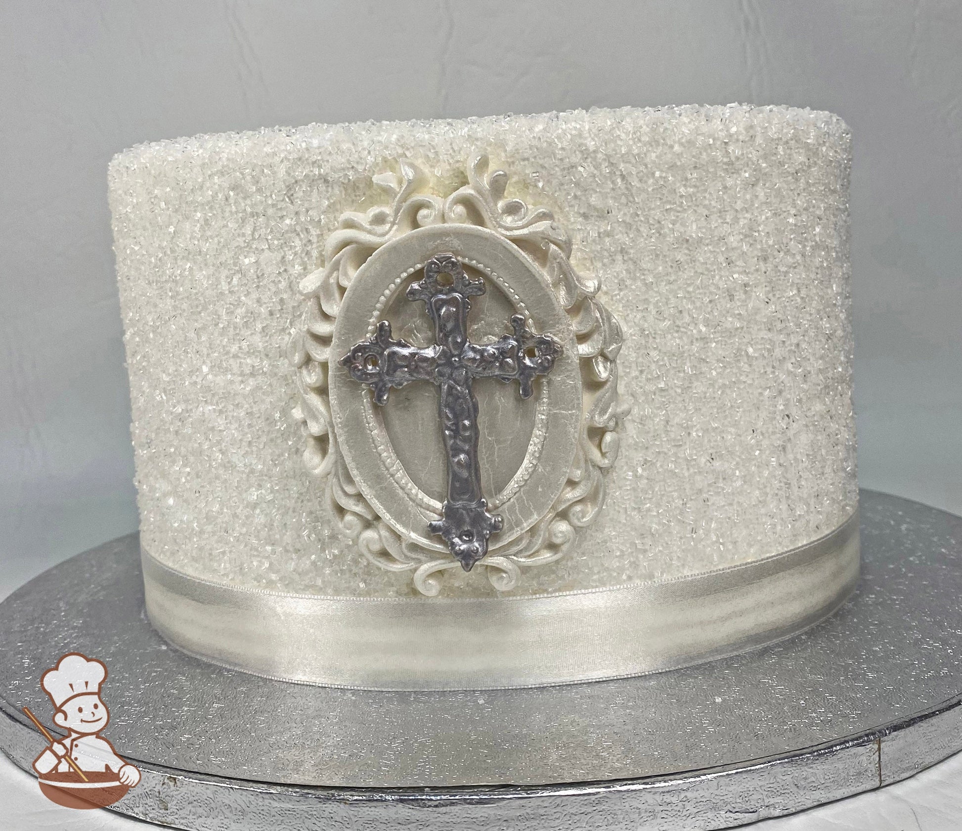 Single tier round cake decorated with white sugar crystal and a silver fondant cross. Lastly, on the base of the cake is a white satin ribbon.