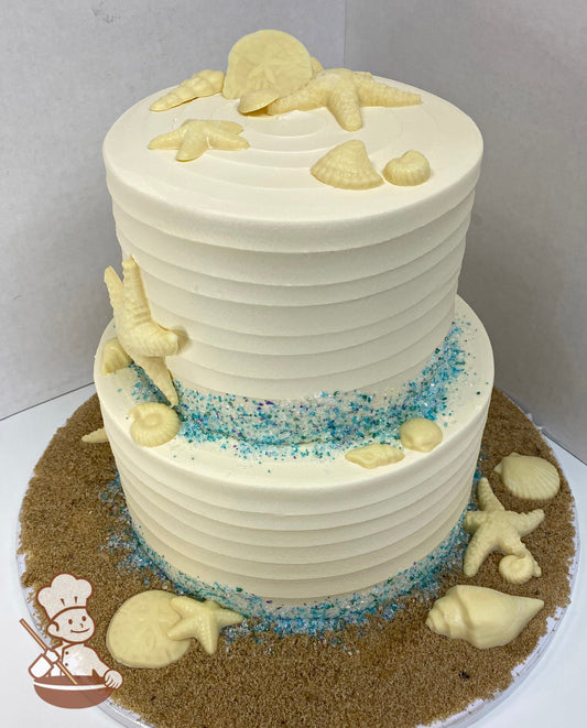 2-tier cake decorated with horizontal textured icing and blue tone sugar crystals on the bases of the tiers. The cake also has edible seashells.