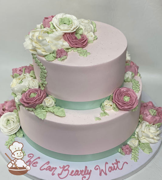 2-tier round cake with pale pink icing and an assortment of buttercream flowers in white and pink colors with light green leaves and vines.