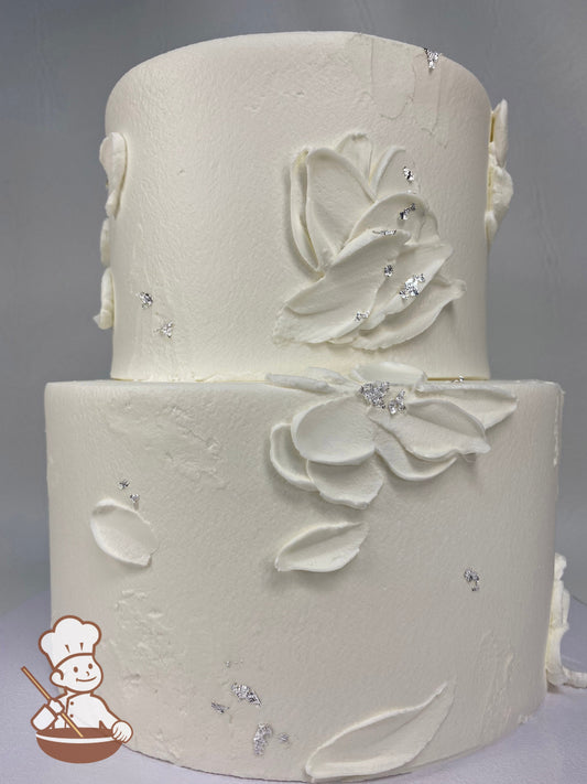 2-tier round cake with white icing and palette knife flowers and a some silver foil flakes on the cake walls.