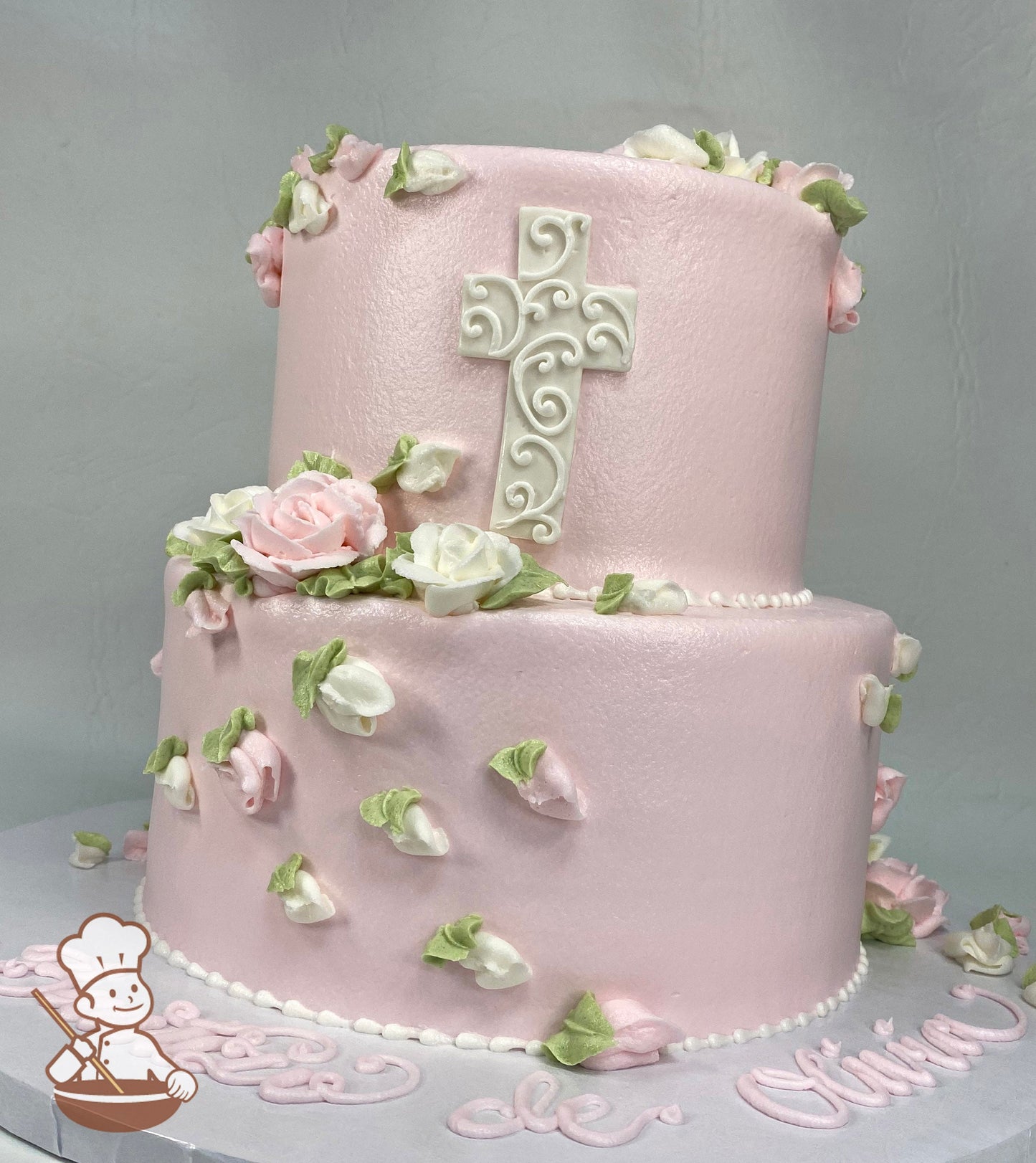 2-tier round cake with smooth light pink icing and buttercream roses in light pink and white colors and a white fondant cross on the cake wall.
