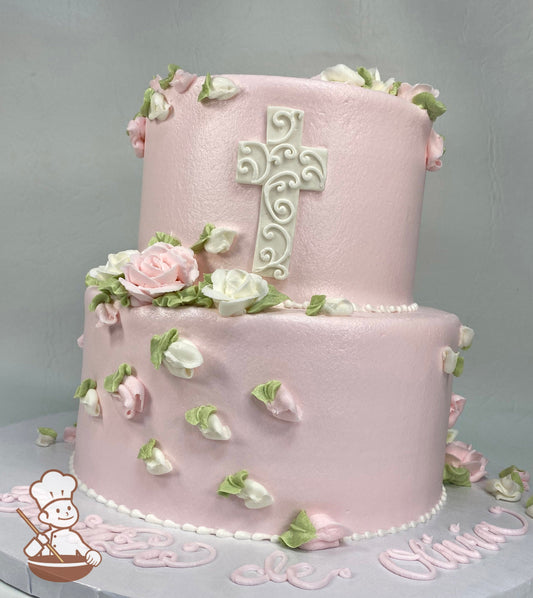 2-tier round cake with smooth light pink icing and buttercream roses in light pink and white colors and a white fondant cross on the cake wall.