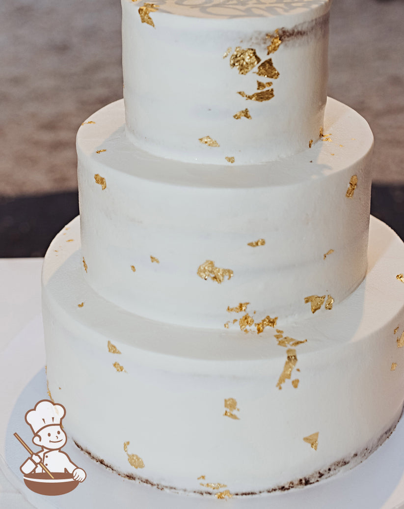 3-tier cake with white icing that has been scraped a bit to show some of the cake underneath and added gold foil flakes on all the tiers.