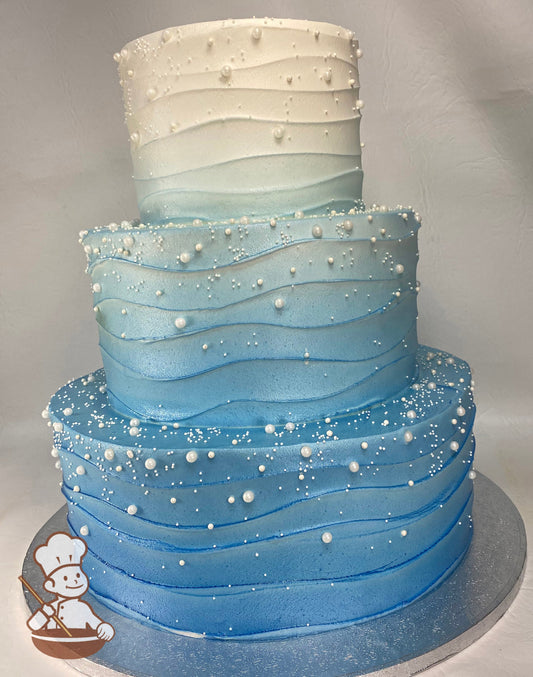 3-tier cake with an Ombre blue icing and a wavy texture, also has white sugar pearls in different sizes scattered all over cake.