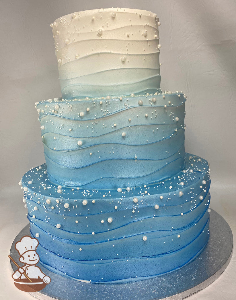 3-tier cake with an Ombre blue icing and a wavy texture, also has white sugar pearls in different sizes scattered all over cake.