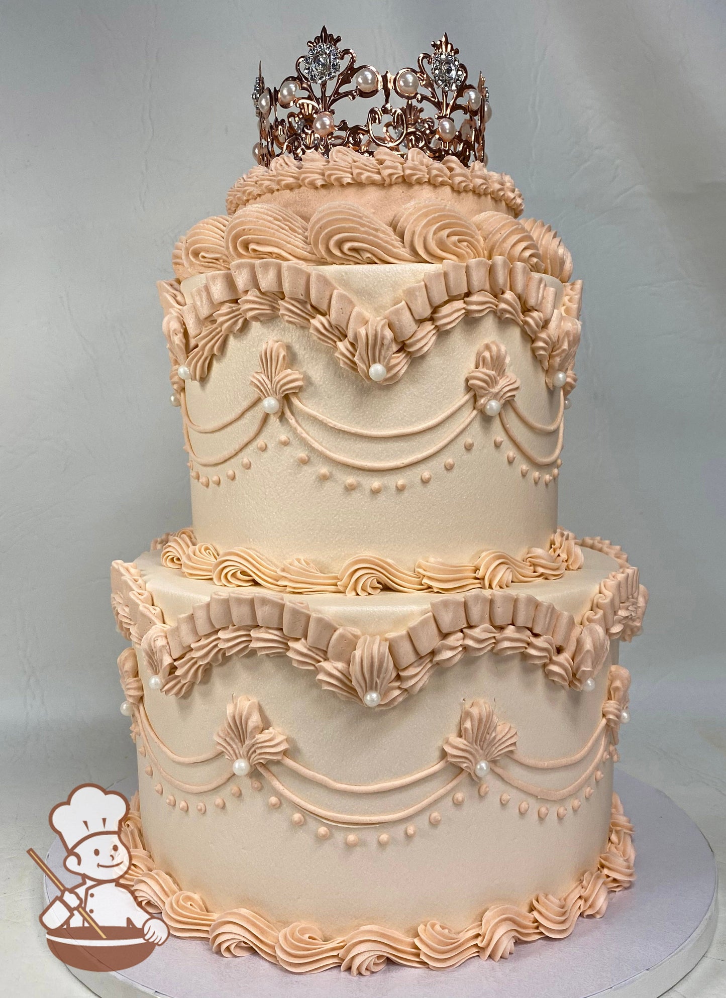 2-tier cake with light peach-colored smooth icing and vintage style piping's with white pearls and a rose gold crown on the top tier.