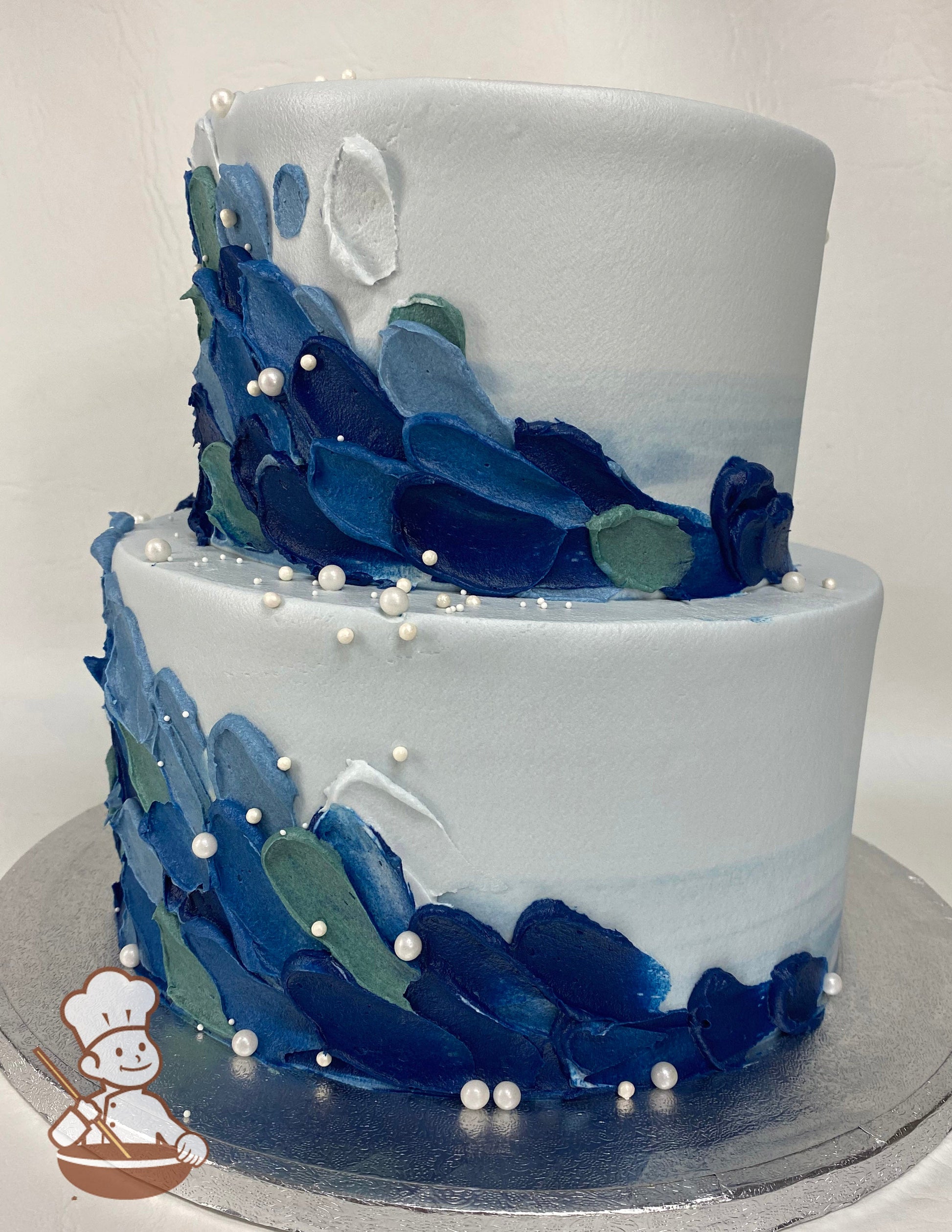 2-tier cake with a light blue watercolor icing, decorated with buttercream palette knife scales in different shades of blue and white pearls.