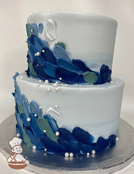 2-tier cake with a light blue watercolor icing, decorated with buttercream palette knife scales in different shades of blue and white pearls.