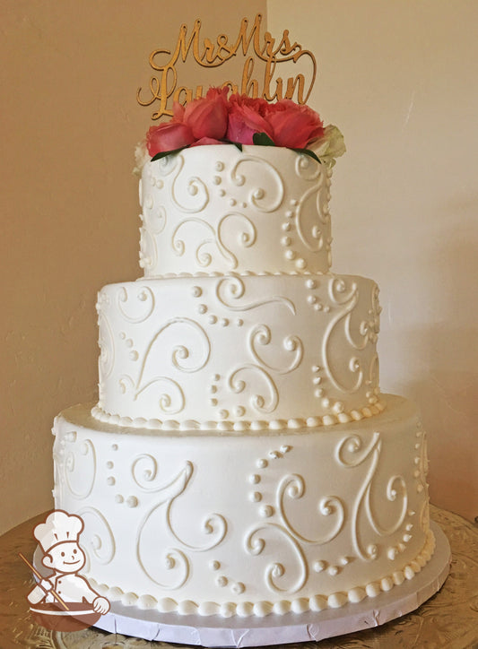 3-tier cake with smooth white icing and white buttercream scrolls and fresh flowers as the topper.