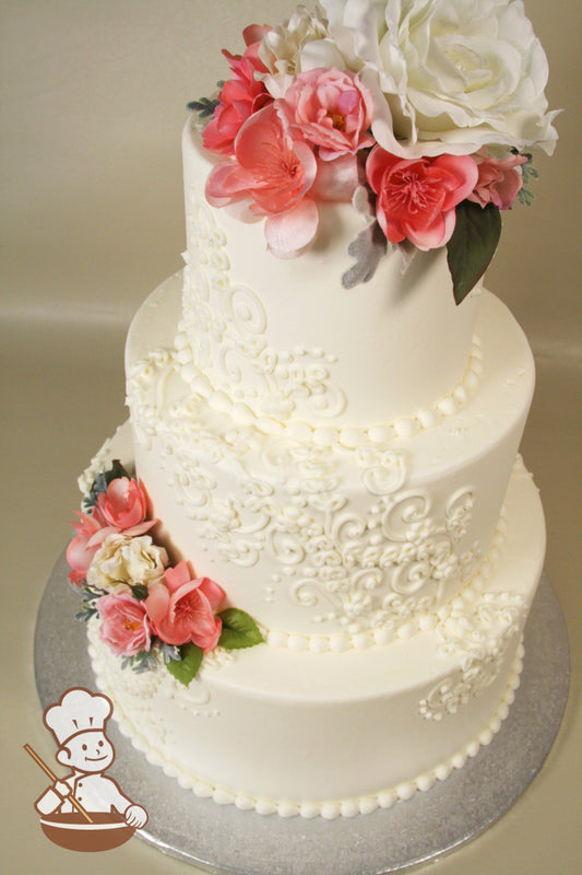 3-tier cake with smooth white icing and decorated with white buttercream scrolls and fresh flowers.