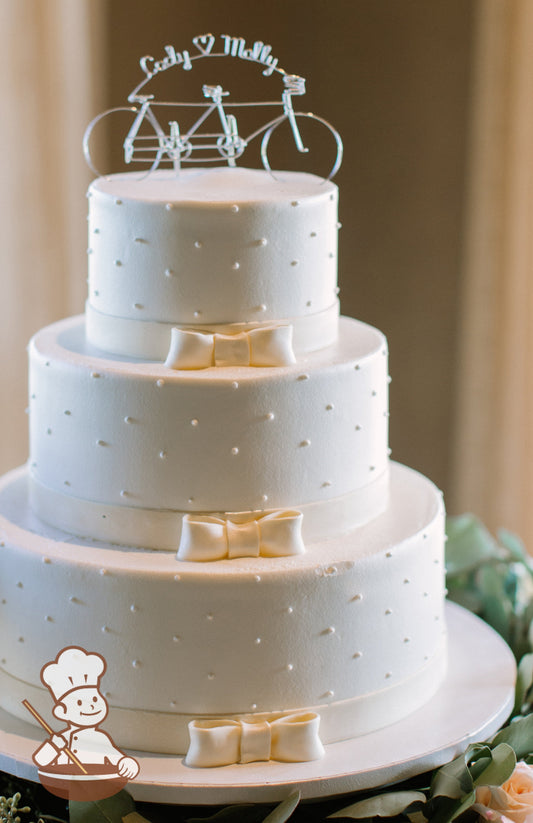 3-tier cake with smooth white icing and decorated with white buttercream dots and a white fondant bow and band.