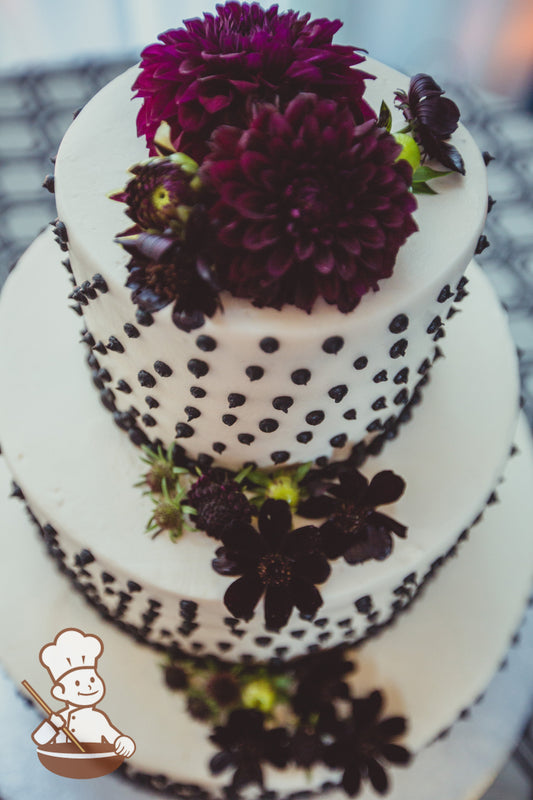 3-tier cake with smooth white icing and decorated with black buttercream dots and fresh flowers.