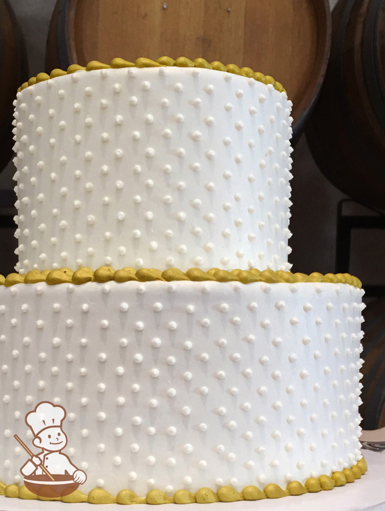2-tier cake with smooth white icing and decorated with white buttercream dots and a yellow-gold beaded trim.