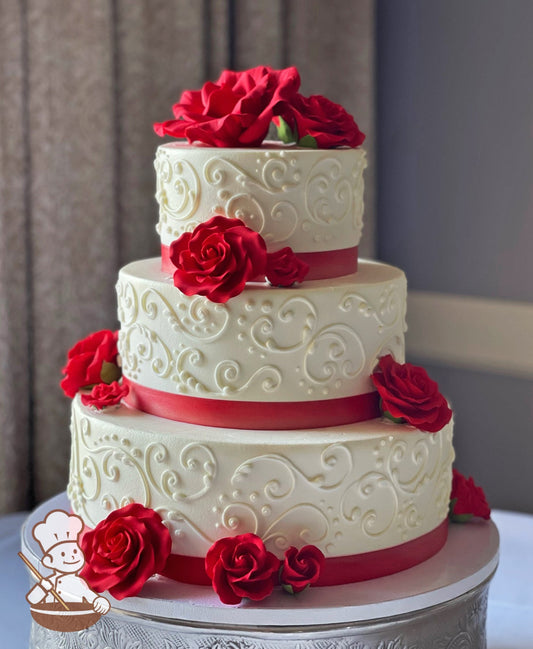 3-tier cake with smooth white icing and decorated with white buttercream scrolls, a red satin ribbon and red sugar roses.