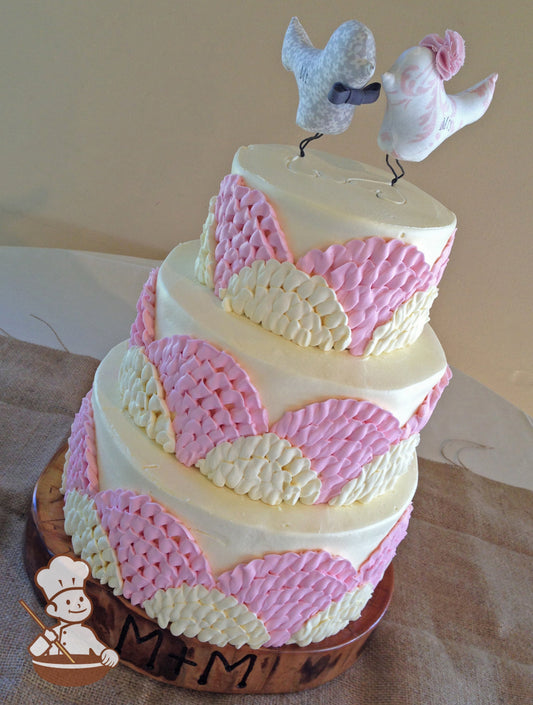 3 tier wedding buttercream cake with pink and white fan shaped pattern and whimsical lovebirds cake topper.