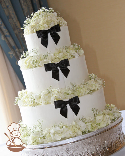 3 tier buttercream wedding cake with tuxedo black satin bow decor and white hydrangeas and baby's breath rings on base of each tier.