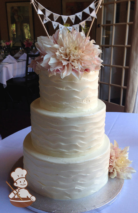 3-tier cake with white icing and decorated with a wavy texture and fresh flowers.