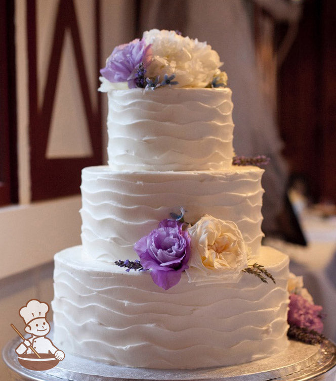 3-tier cake with white icing and decorated with a wavy texture and fresh flowers in white and purple colors.