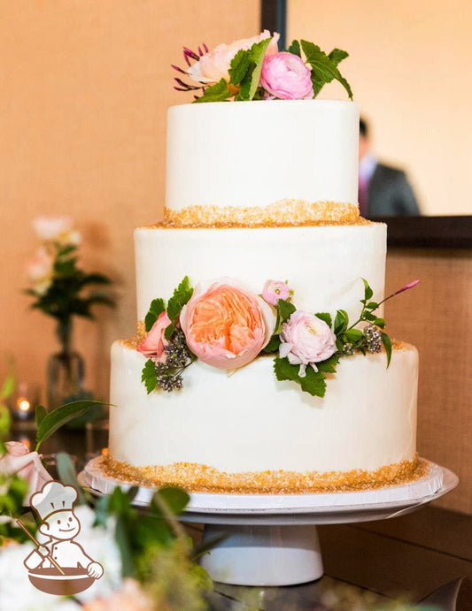 3-tier cake with smooth white icing and decorated with gold sugar sand on the bases of the tiers and fresh flowers.