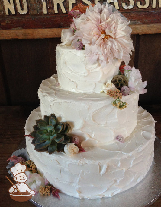 3-tier cake with white icing and decorated with prominent texture all over the cake and fresh flowers.