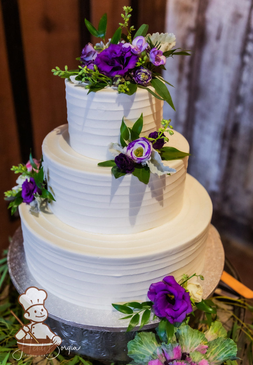 3-tier cake with white icing and decorated with a horizontal texture and fresh flowers in purple colors.