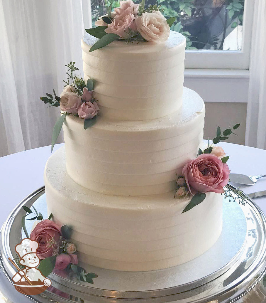 3-tier cake with white icing and decorated with a light horizontal texture and fresh flowers in different shades of pink.