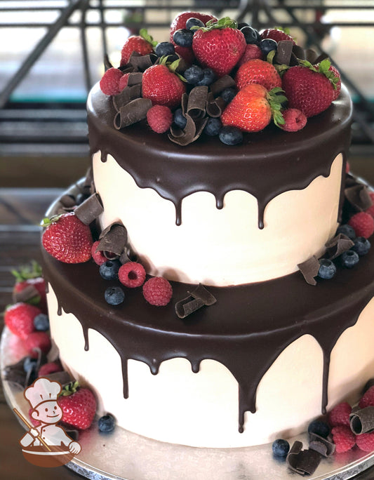Cake with white smooth icing and decorated with a chocolate ganache pour, fresh strawberries, raspberries, blueberries and chocolate curls.