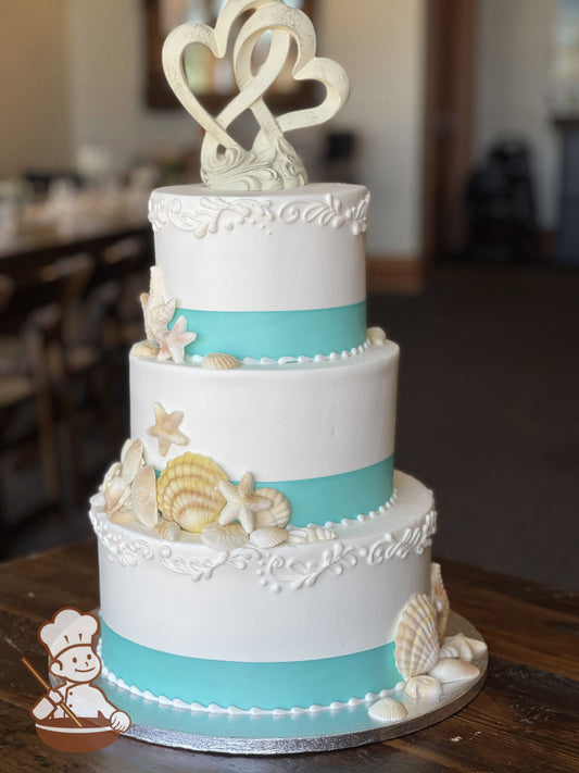 3-tier cake with thin scroll pattern piped on the top and bottom tier. All tiers with baby blue ribbon and decorated with sea shells.