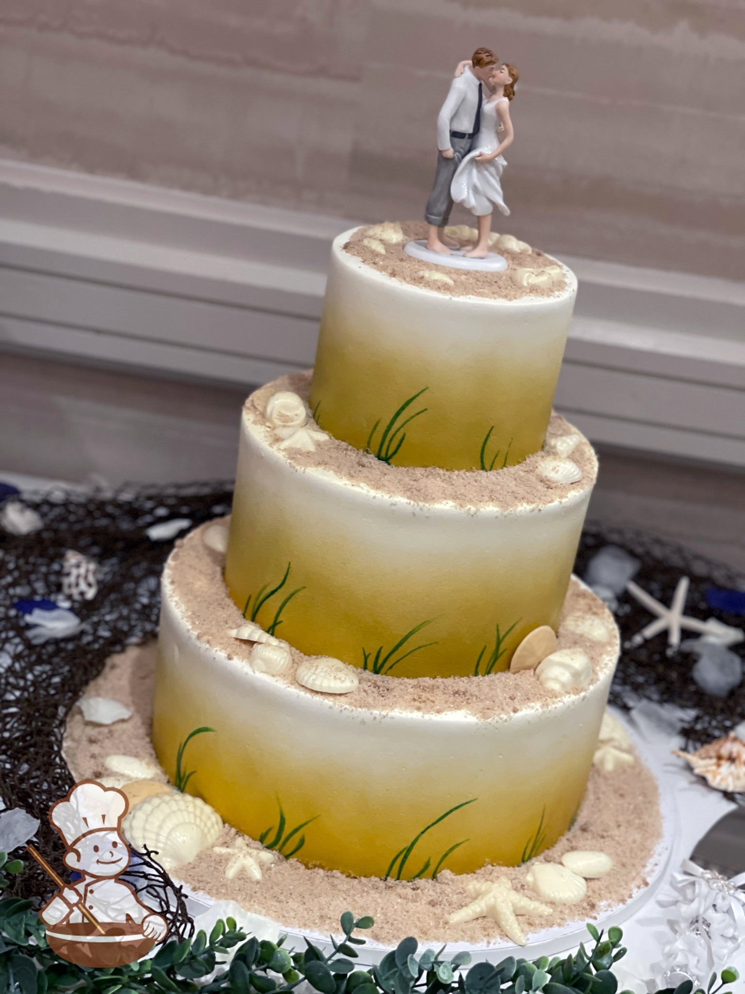 3-tier cake with brown sugar sand on top of each tier with sea shells. Cake wall are sprayed sandy brown and hand piped green blowing grass.