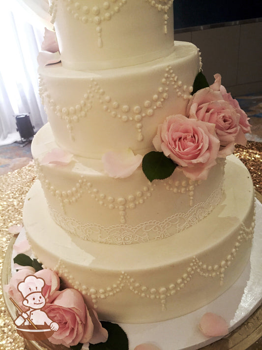 4-tier cake with smooth white icing and white buttercream piping's and a sugar lace trim on one tier and light-pink fresh roses.