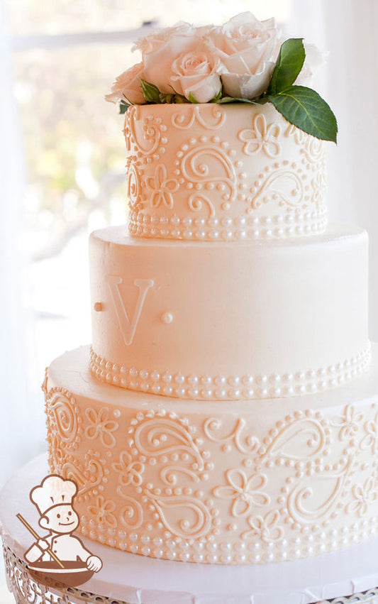 3-tier cake with ivory-tinted smooth icing and decorated with buttercream piped flowers and scrolls on the bottom and top tier and a monogram.