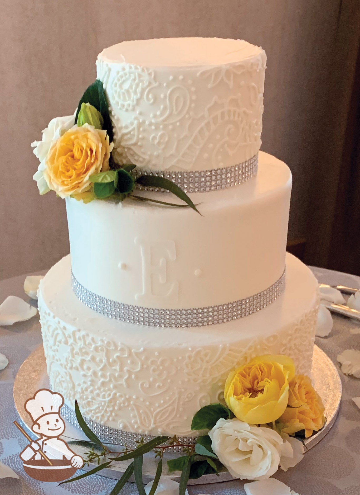 3-tier cake with smooth white icing and decorated with buttercream piped flowers and scrolls on bottom and top tier and a fondant monogram.