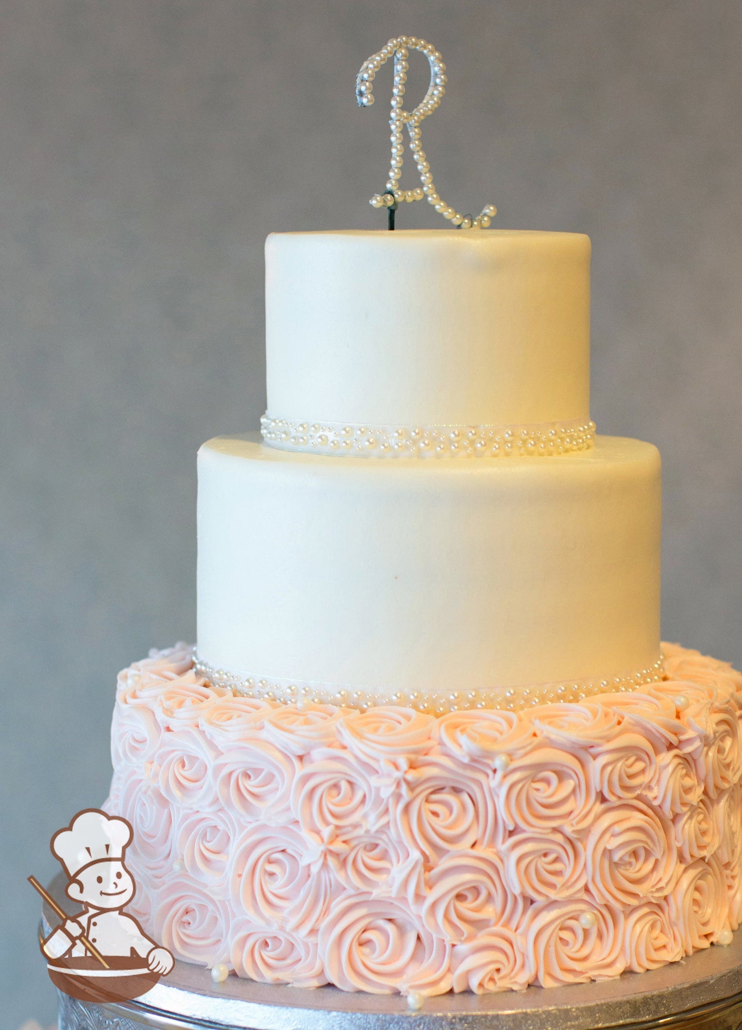Light pink rosette swirls on bottom of a 3 tier round cake and smooth buttercream iced top tiers with white pearl bead band.