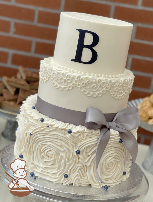 3 tier buttercream wedding cake with large white floral swirl & piping finished with monogram, satin ribbon bow and navy blue pearl beads.