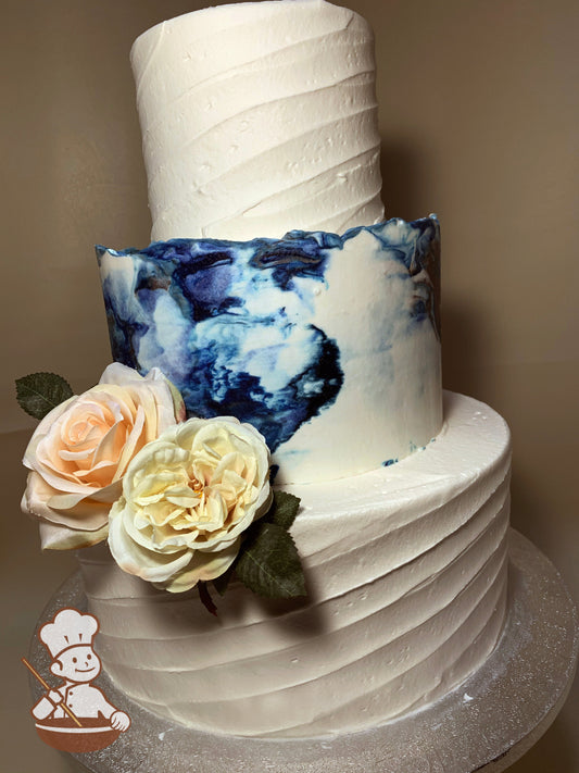 3 tier wedding cake with dark & light blues marble buttercream in middle and diagonal texture for bottom and top tiers.  Pastel flowers added.