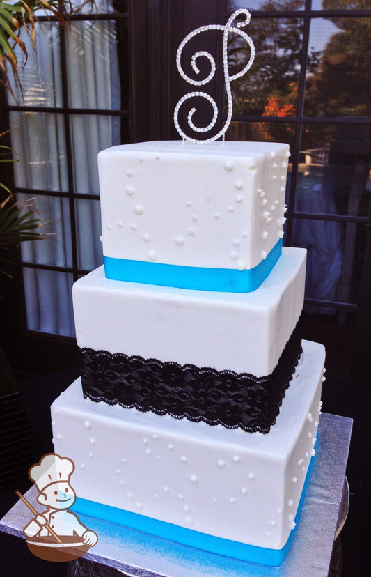 3 tier square wedding cake with black fabric lace wrapped middle tier and blue satin ribbon wrapped other tiers and finished with beaded piping.