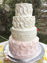 4-tier cake decorated with light-pink rosettes on the bottom tier, white scrolls and pearls on the second tier and texture on third and top tier.