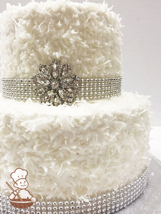 2-tier cake with coconut flakes pressed all over the cake and a rhinestone band on the base of each tier with a brooch on the top tier.