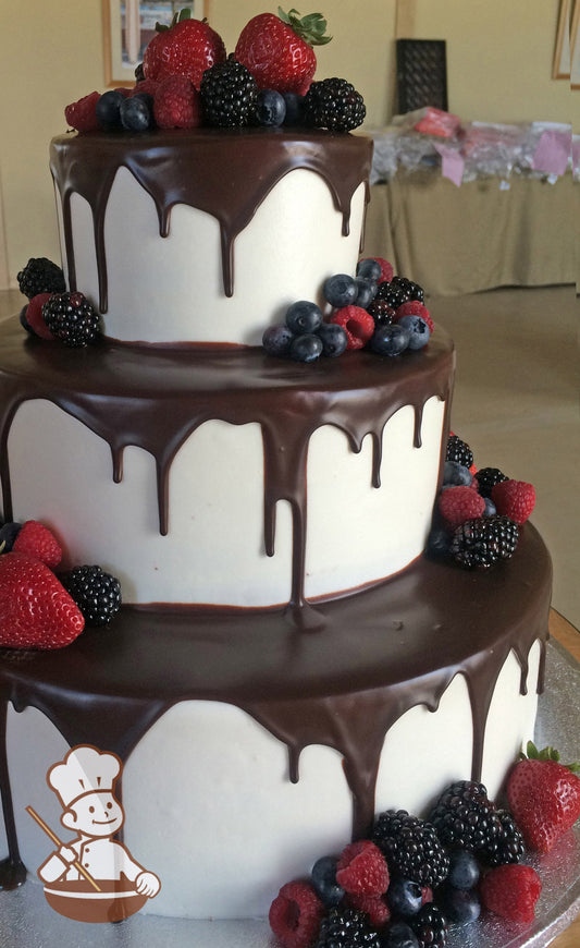 3-tier cake with smooth white icing and decorated with a chocolate ganache pour and fresh strawberries, raspberries, blackberries & blueberries.