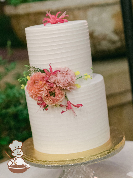 2-tier cake with white icing and decorated with thin horizontal texture and fresh flowers.