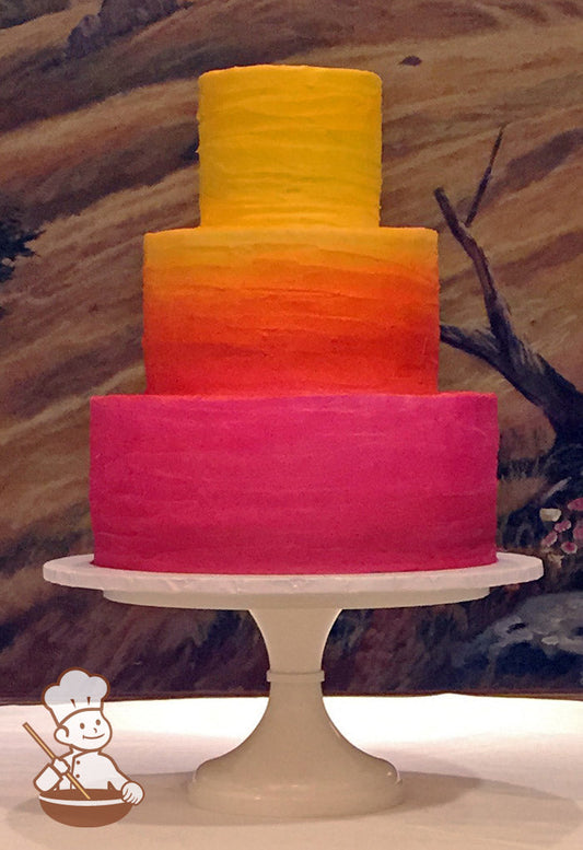 3-tier cake with white icing that has been decorated with a messy horizontal texture and an airbrushed ombre spray in sunset colors.