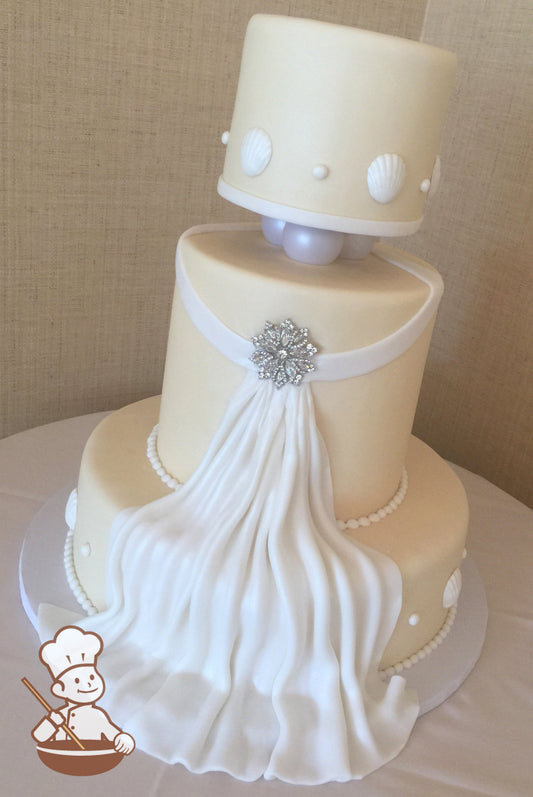 3 tier ivory fondant wedding cake with fondant fabric flowing down the cake & a crystal broach center and finished with seashells & pearl beads.