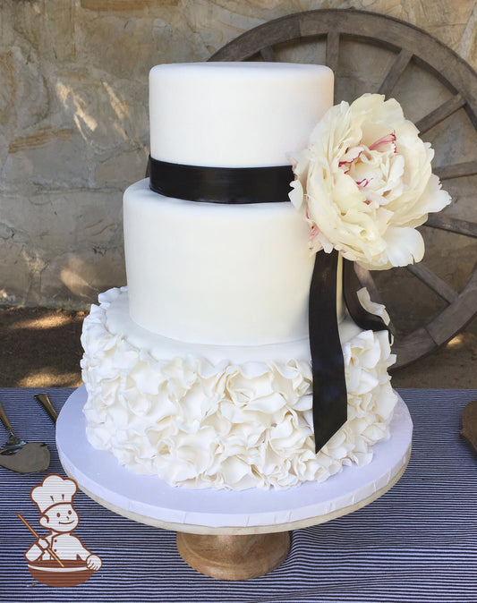3 tier wedding cake with fondant petal ruffle wrap on bottom tier decorated with black fondant ribbon and fresh flowers.