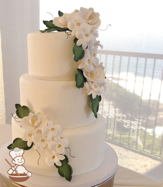 3 tier white fondant wedding cake with cascading white sugar flowers and green sugar leaves.