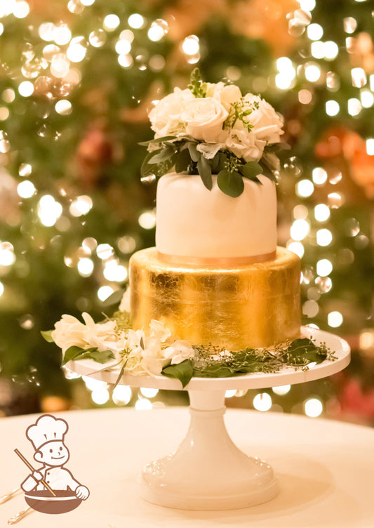2-tier cake with the bottom tier fully covered in gold and the top tier smooth white icing with white fresh flowers.