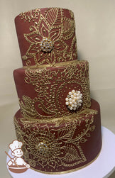 3-tier brick red cake with metallic gold piping of Indian themed scrolls and flowers, with diamond and pearl broche in the piped flowers.
