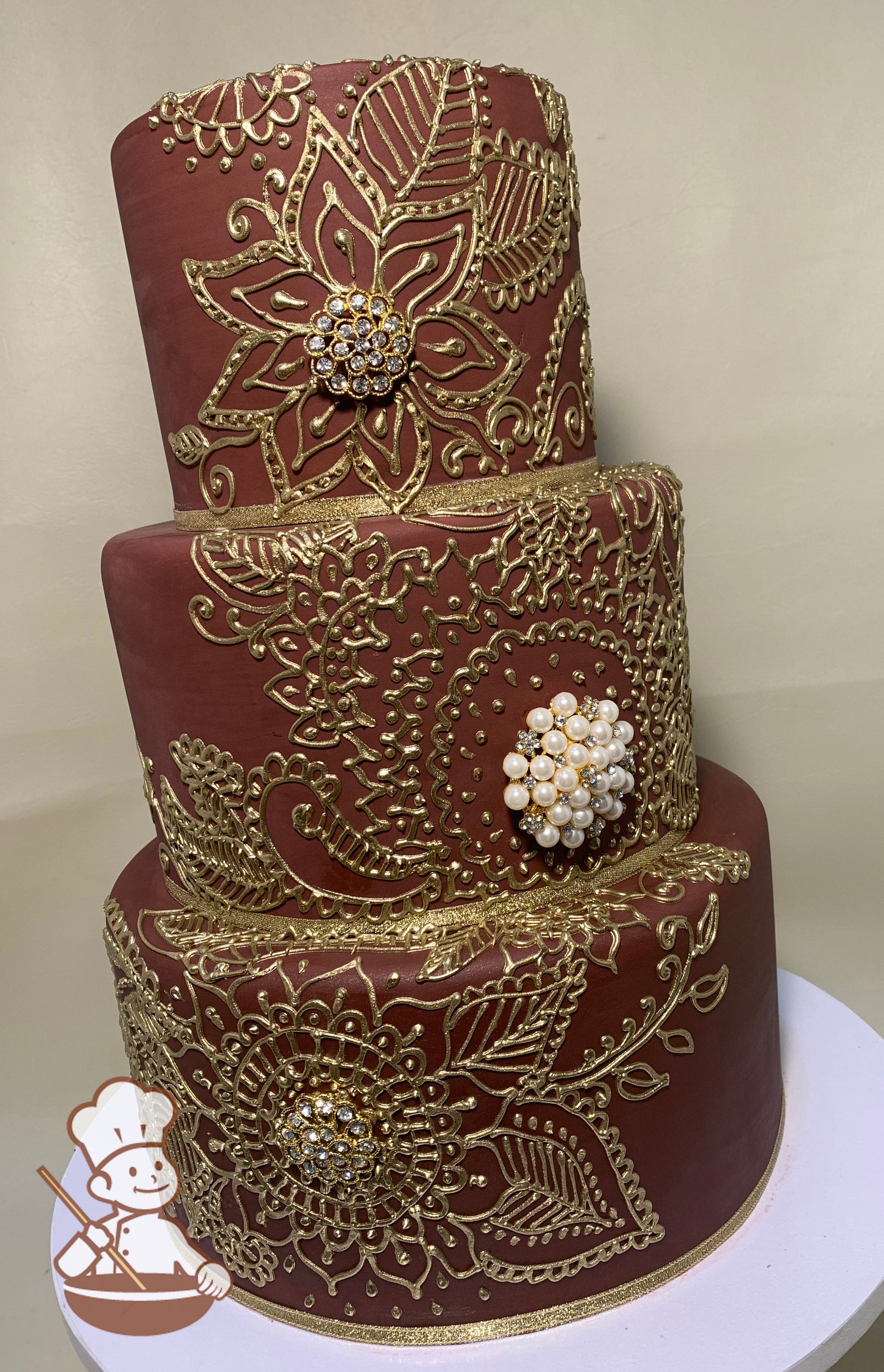 3-tier brick red cake with metallic gold piping of Indian themed scrolls and flowers, with diamond and pearl broche in the piped flowers.